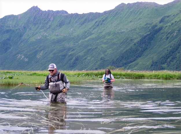Photo of Dr. Andrew Skibo and Kate Mohatt wading through water and applying chemicals to the water. Mountains in the background.